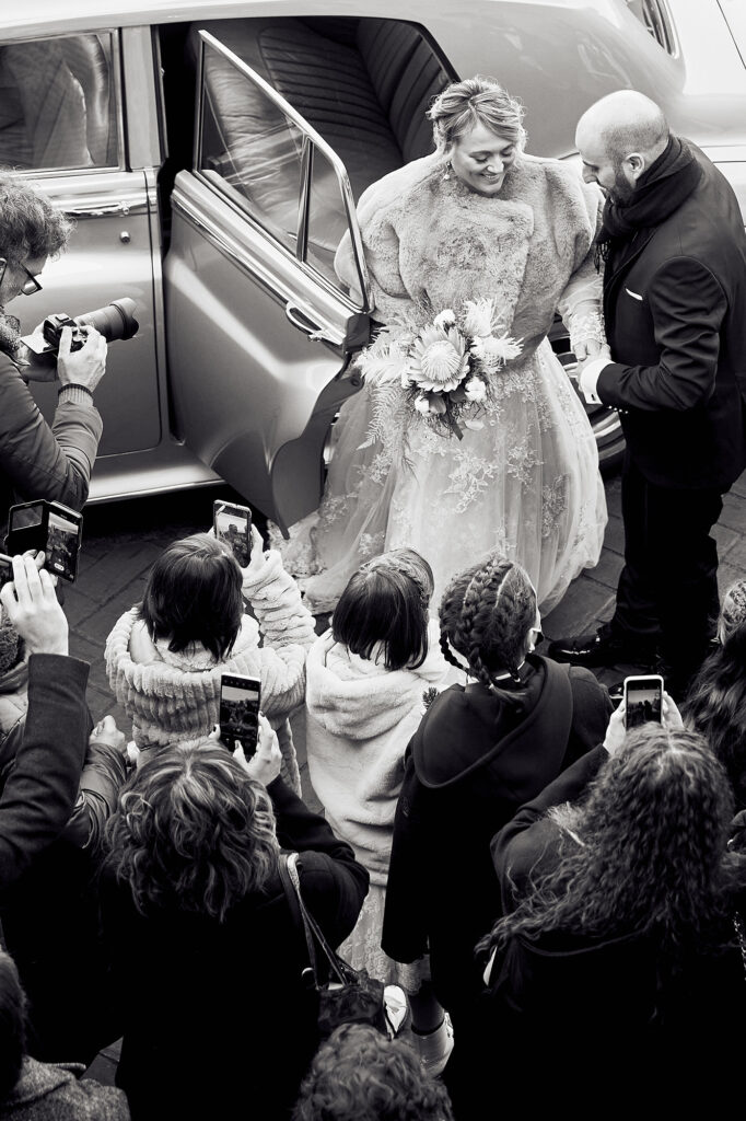 Bride stepping out of the wedding car surrounded by guests